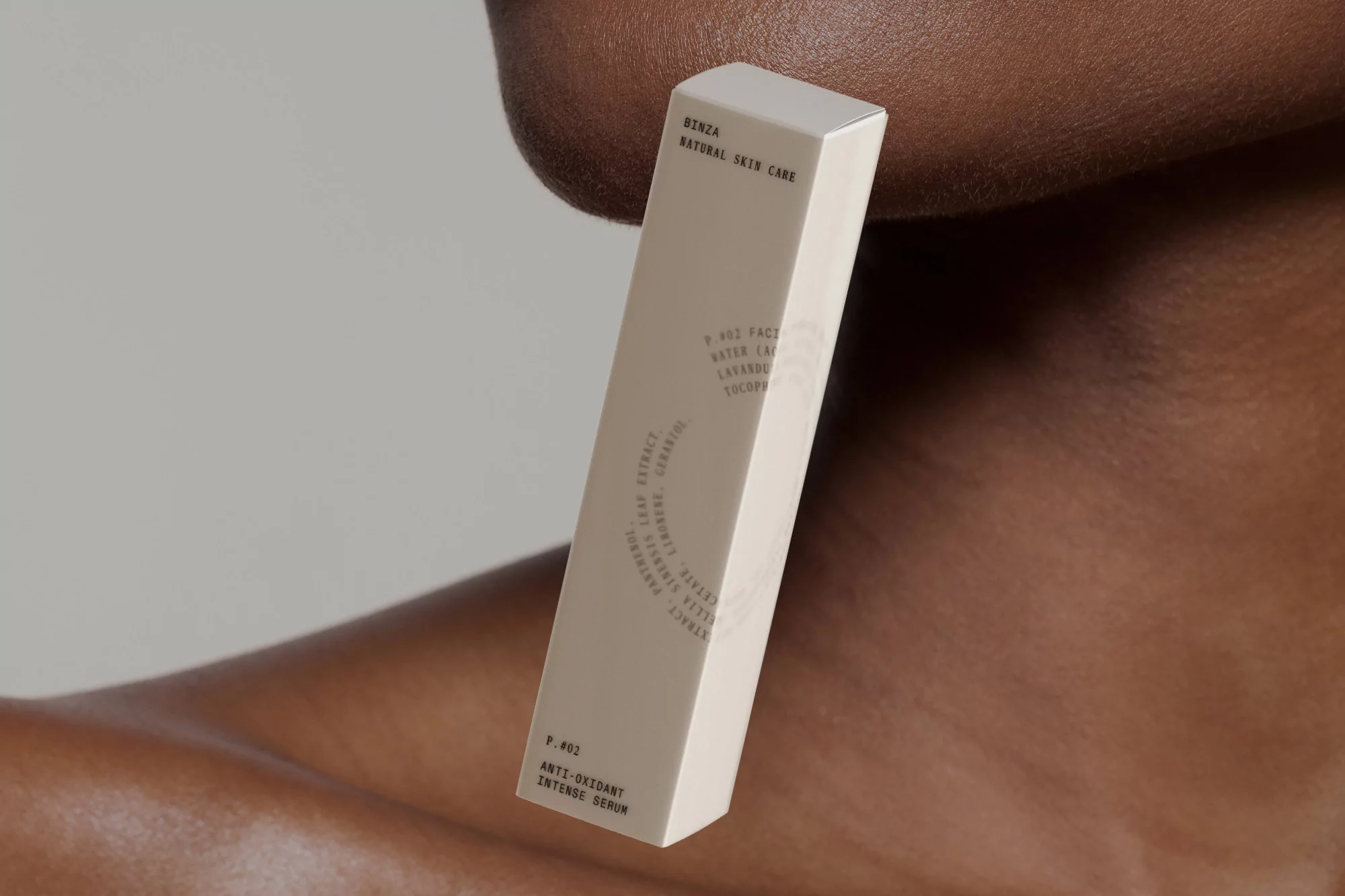 Tan serum box featuring Binza Natural Skin Care brand, with text details, set against a dark-skinned person and light gray backdrop.