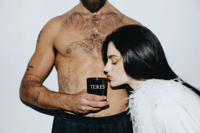 Tattooed, shirtless man holds a black cup labeled 'TEKES' while a woman in a feathered garment kisses the cup. Studio setting.