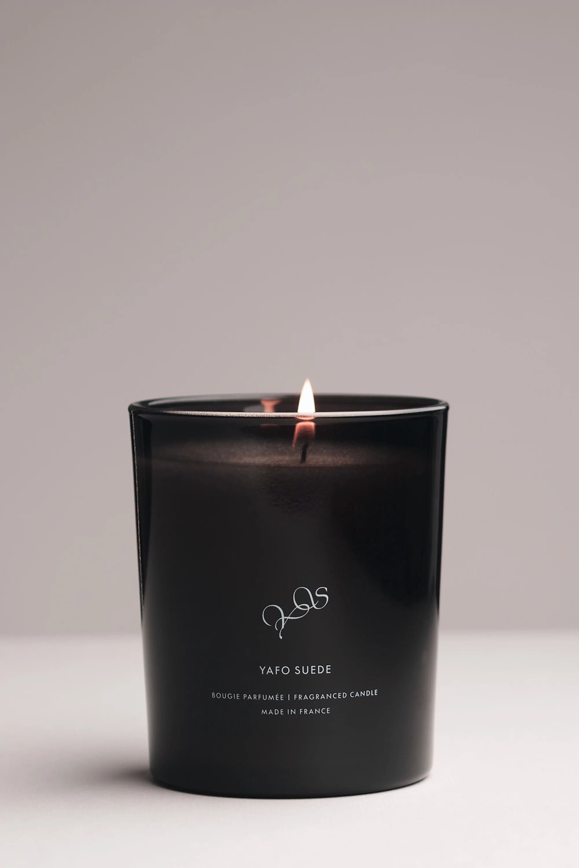 Illuminated YAFO SUEDE candle in a dark container on a white surface with a light gray background.