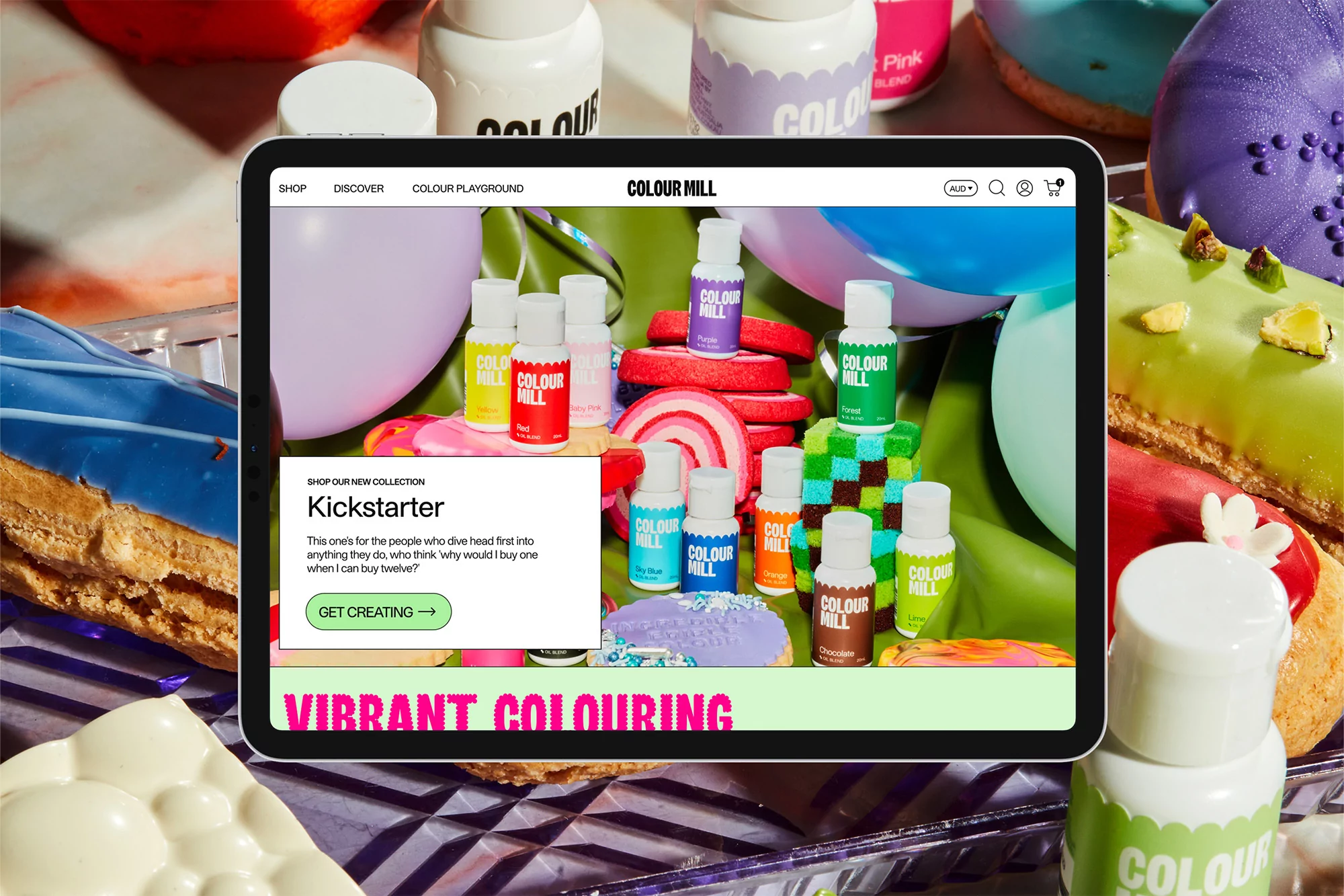 Tablet displaying the Colour Mill website with colorful frosting and food coloring, surrounded by donuts and dessert ingredients.