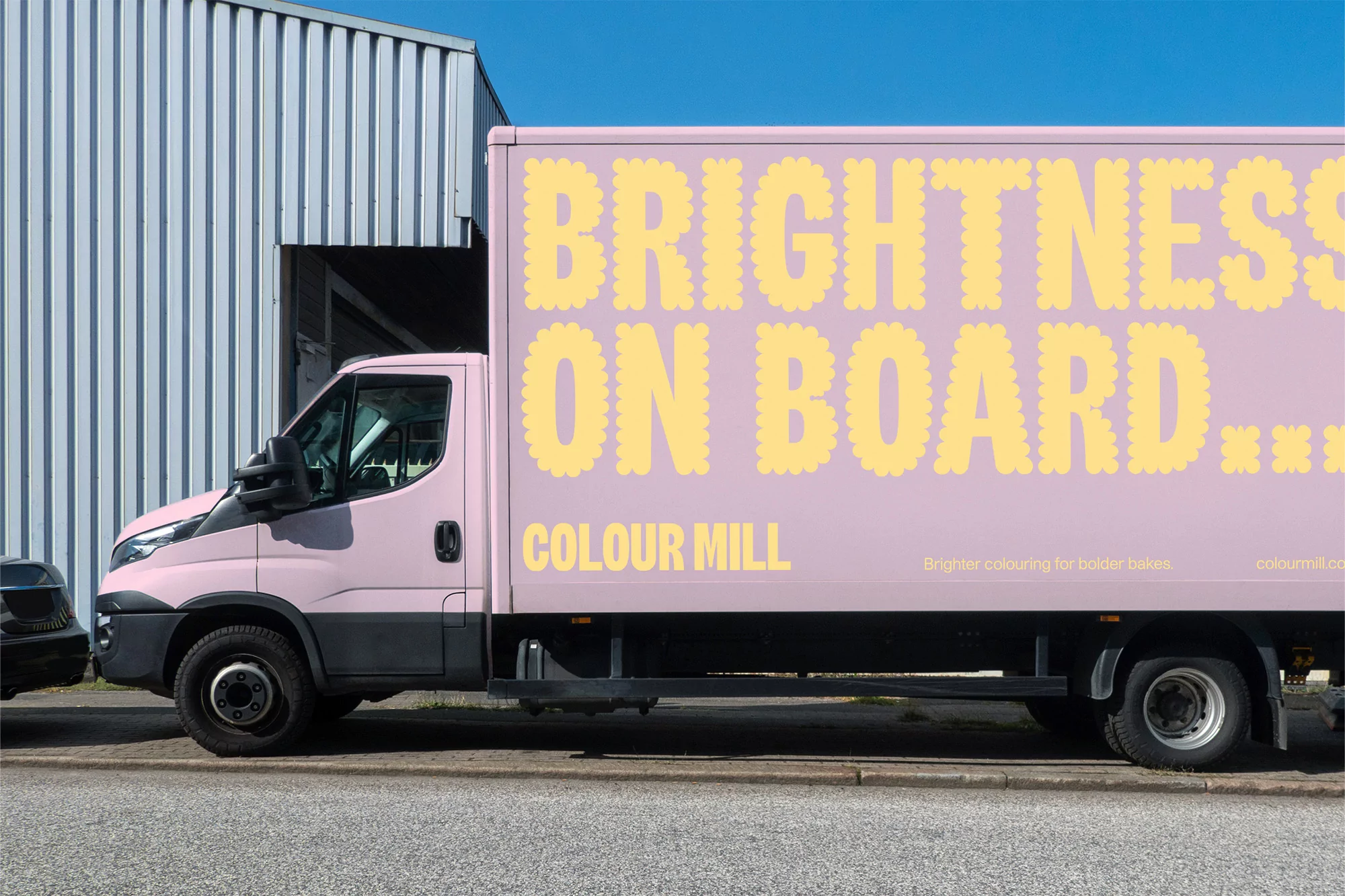 Pink commercial delivery truck with yellow “Brightness on Board…” text on its side, parked on a city street beside a metal building.