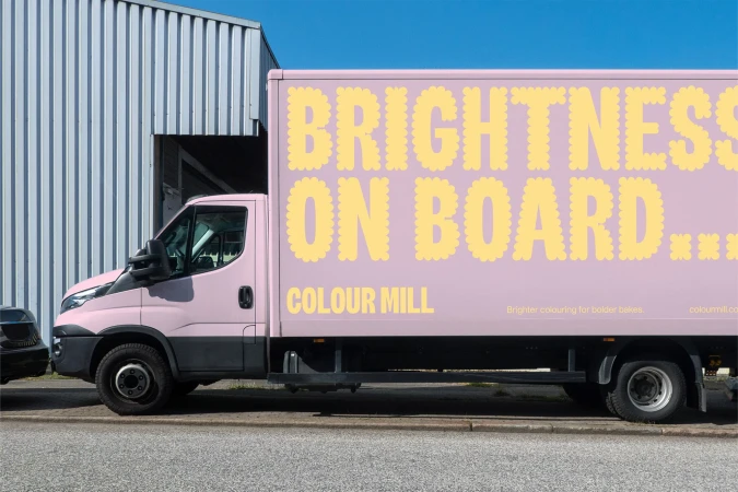 Pink commercial delivery truck with yellow “Brightness on Board…” text on its side, parked on a city street beside a metal building.