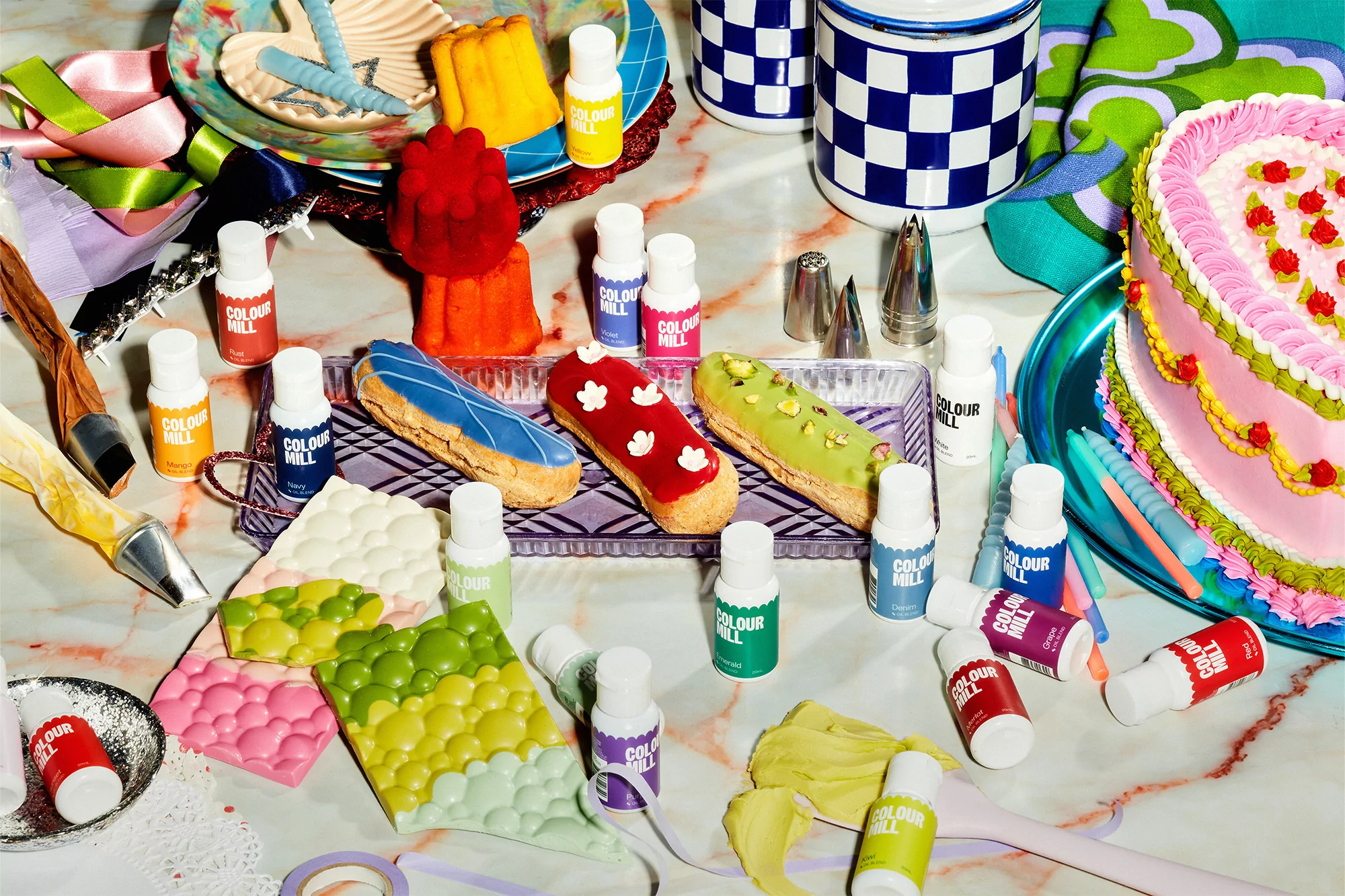 Colorful baking scene featuring éclairs, jello molds, a tiered cake, chocolate bars, and a variety of Colour Mill food colorings.
