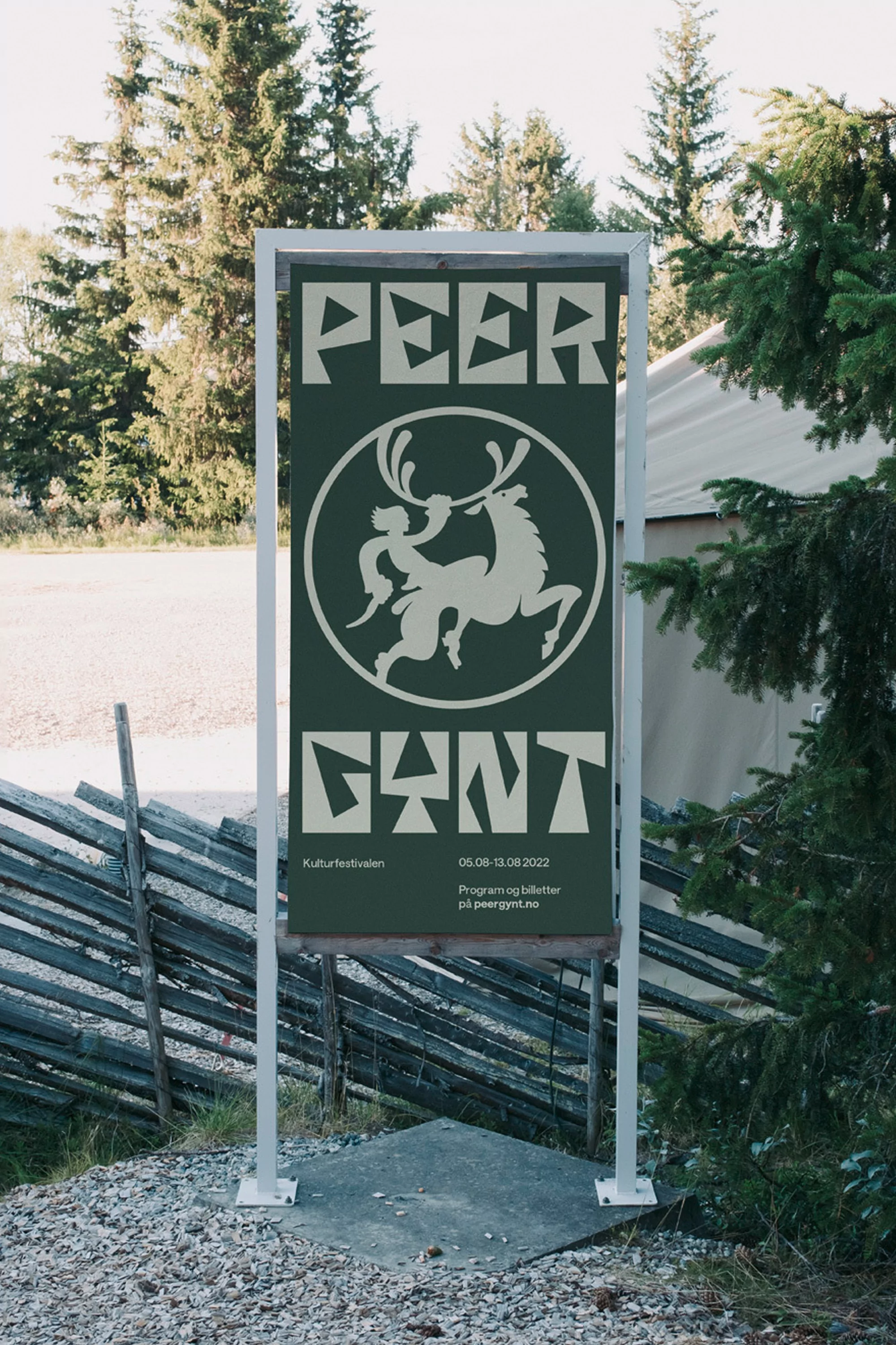Outdoor sign featuring 'Peer Gynt' artwork and text, framed by greenery and a rustic wooden fence in a rural setting on a sunny day.