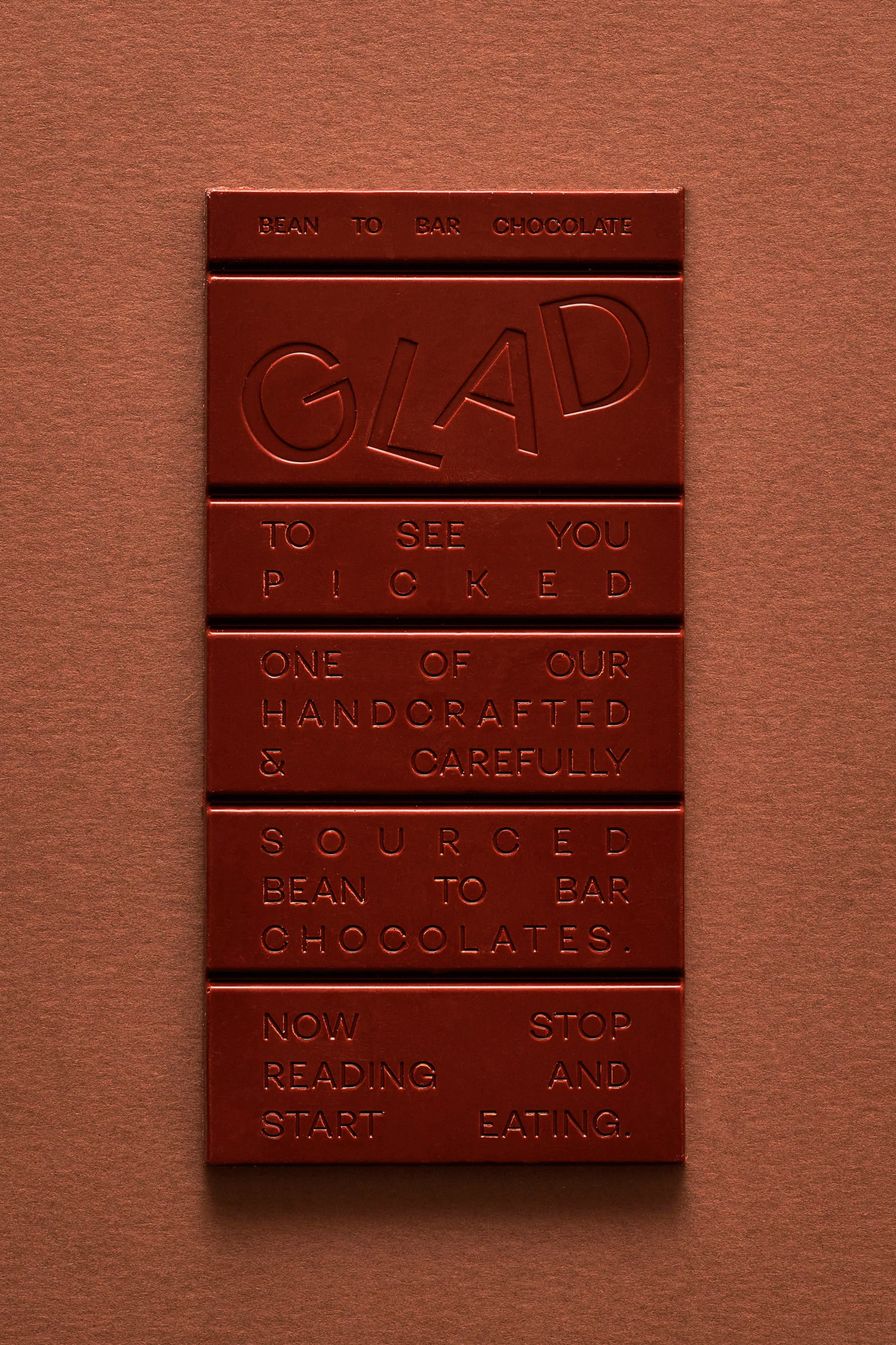 Close-up of a red-brown chocolate bar with embossed text on a matching background. The words 'Glad' and 'Bean to Bar Chocolate' are visible.