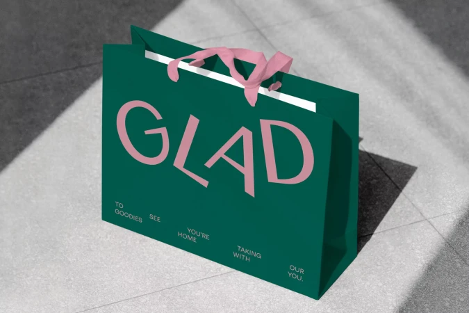 A teal shopping bag with a pink "GLAD" logo sits on a gray tiled floor with soft shadow patterns. The bag has pink ribbon handles.