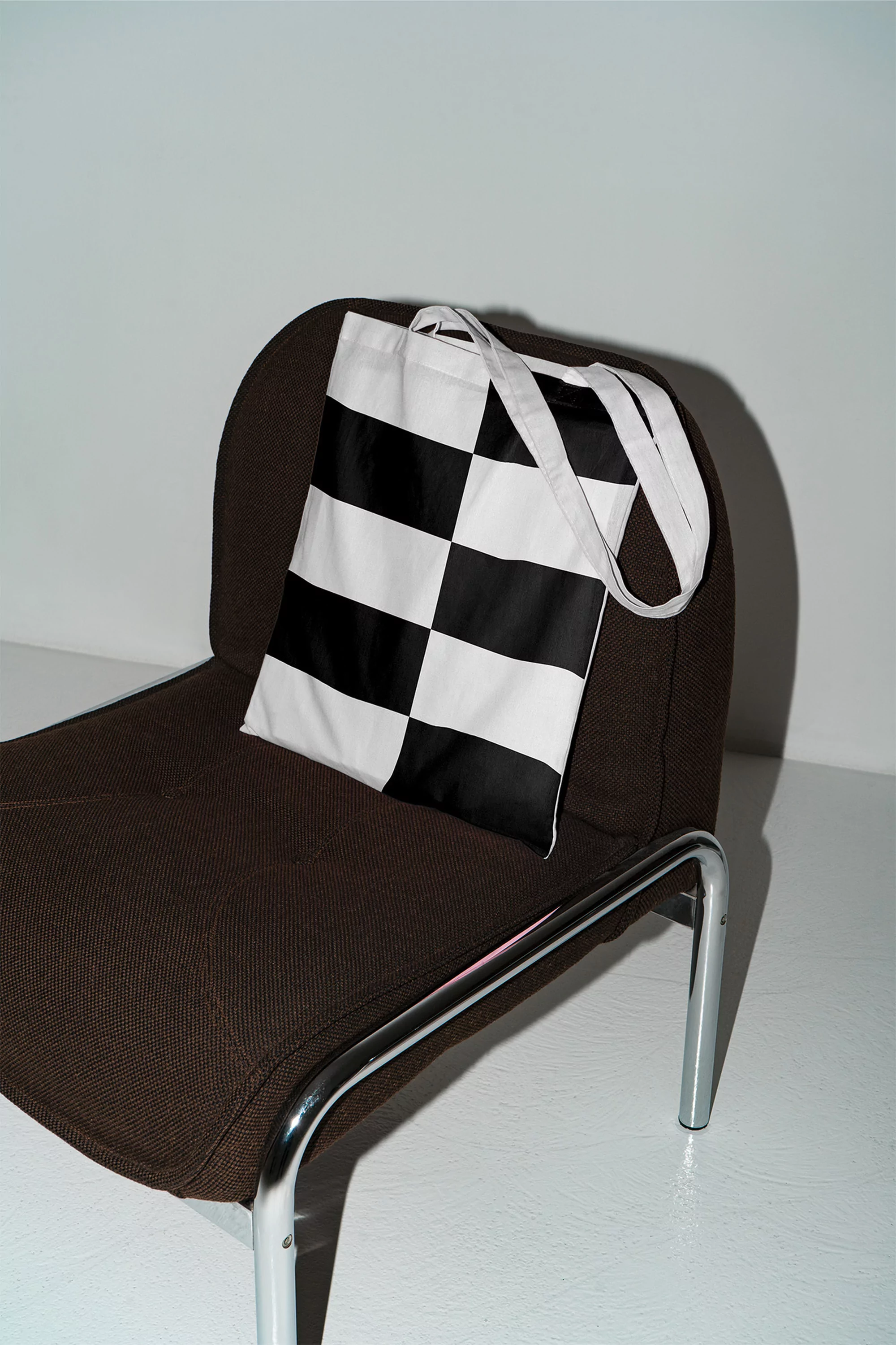 Black and white checkered tote bag resting on a brown chair against a bright white background in a minimalist setting.