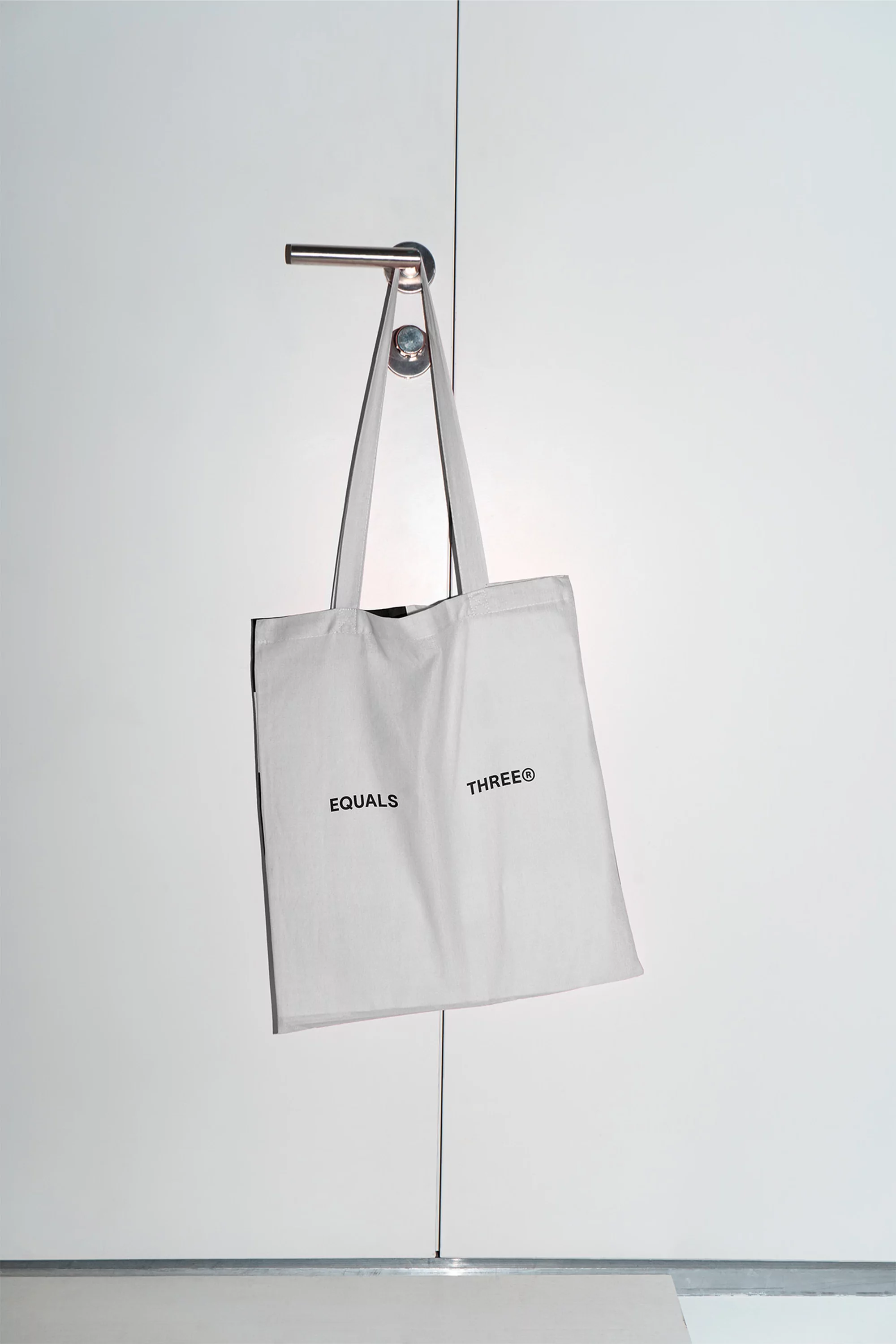 White tote bag hangs from a silver door handle. The words 'Equals Three' are printed on the bag. White background with grey baseboard.