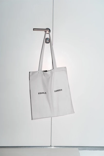 White tote bag hangs from a silver door handle. The words 'Equals Three' are printed on the bag. White background with grey baseboard.