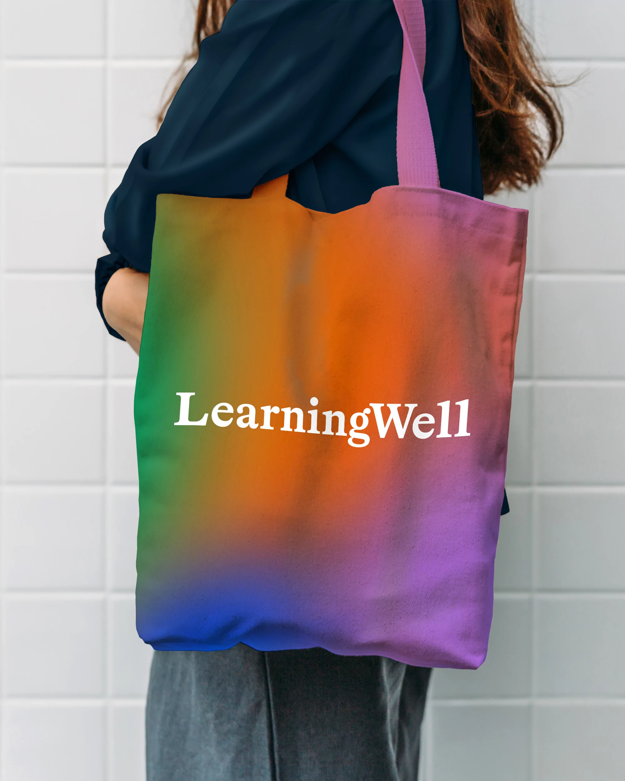 Person with an ombre-colored tote bag over their shoulder with the text "LearningWell" against a minimalist white tiled wall indoors.