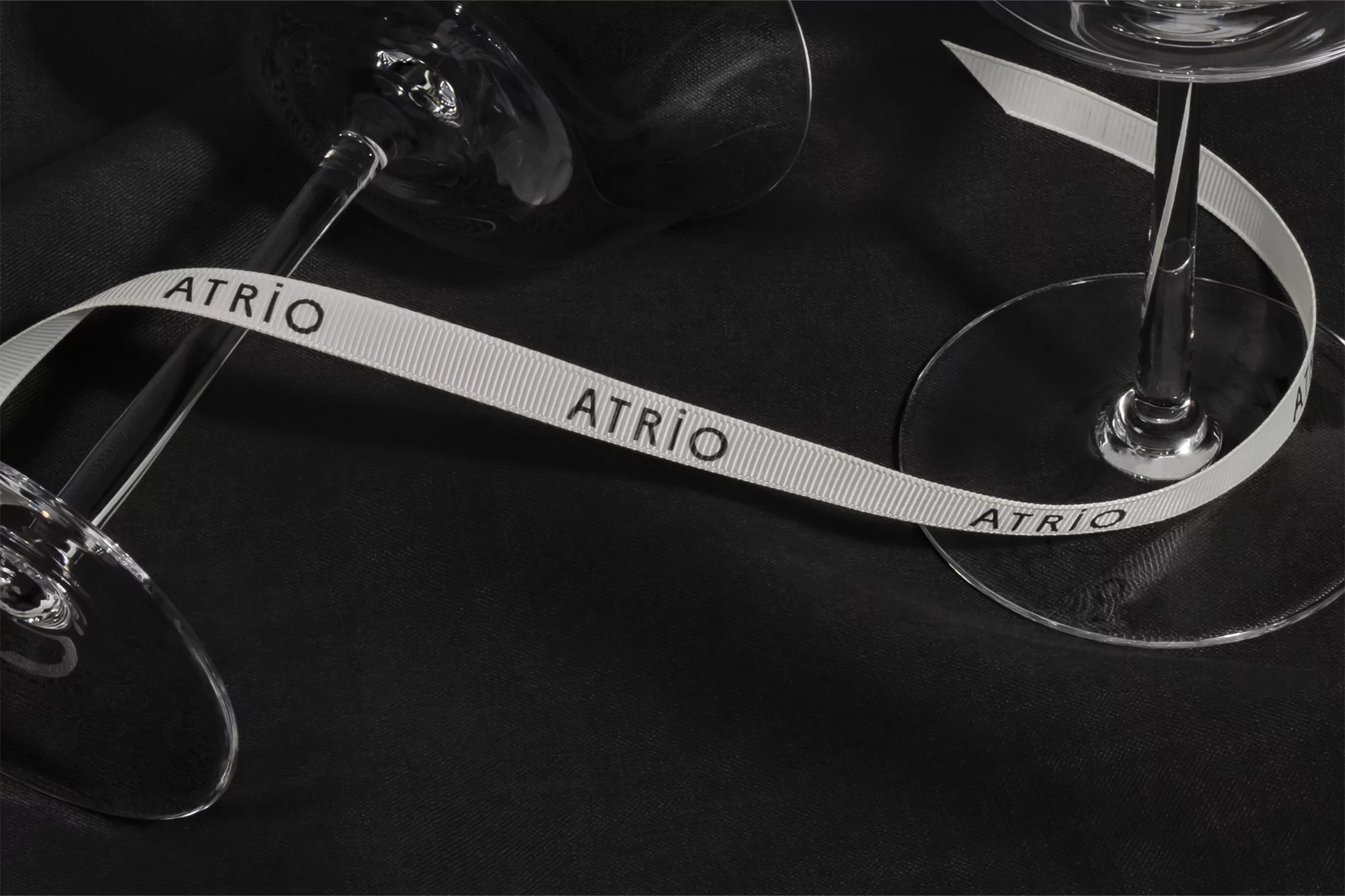 Elegant product shot featuring two wine glasses partially visible, connected by a white ribbon with the text "ATRIO" on a dark surface.