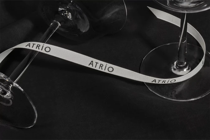Elegant product shot featuring two wine glasses partially visible, connected by a white ribbon with the text "ATRIO" on a dark surface.