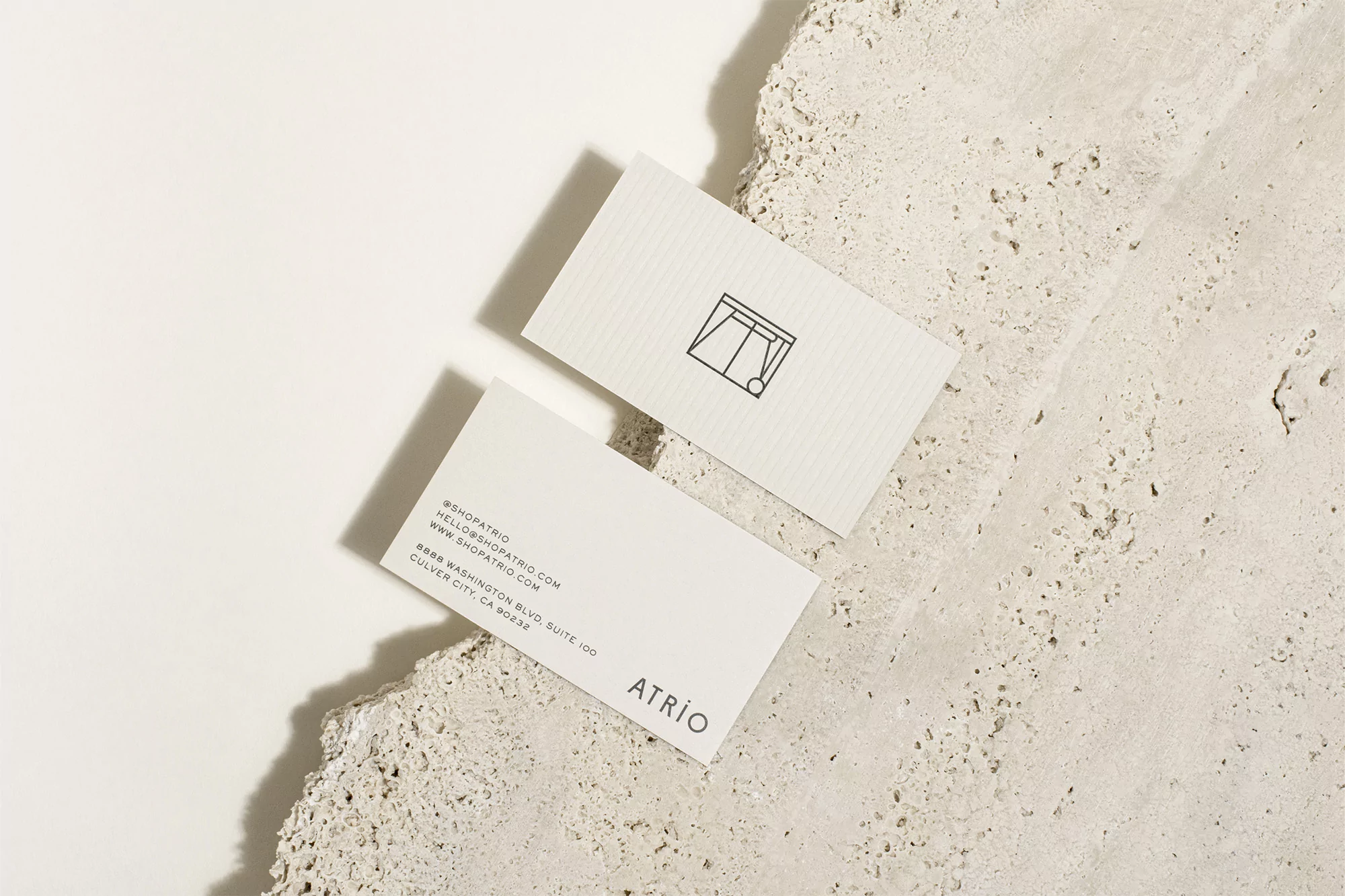 Two business cards featuring a minimalist design and 'ATRIO' logo, resting on a textured stone surface with soft, natural lighting.