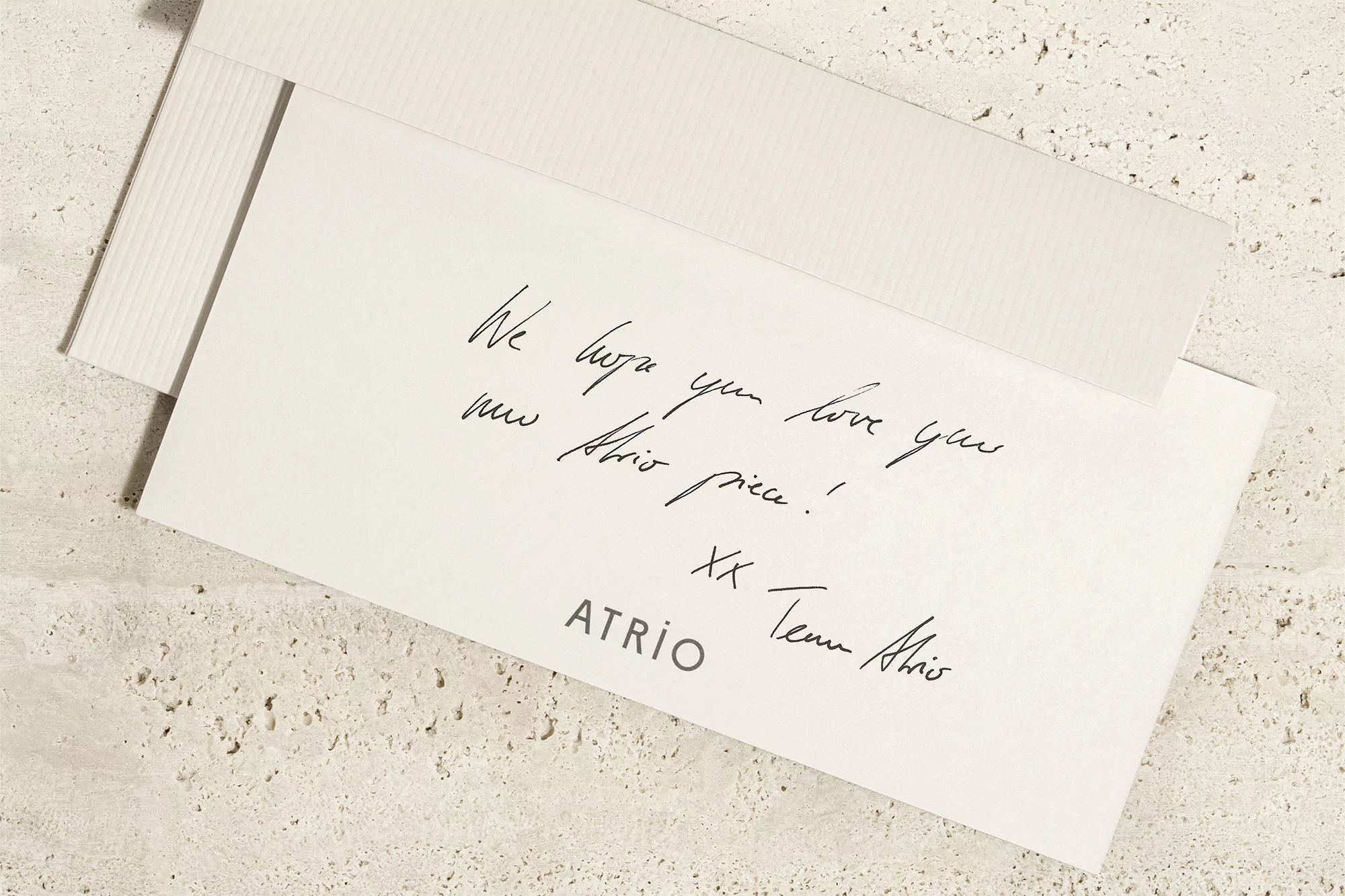 Close-up shot of a white greeting card with handwritten text and the brand name ATRiO on a textured beige background, soft lighting.