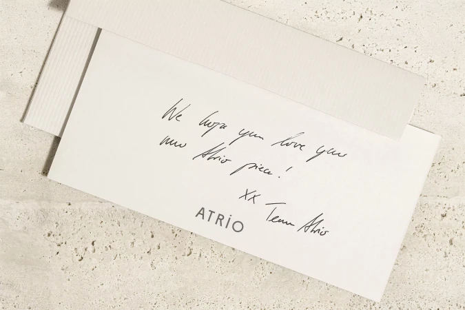 Close-up shot of a white greeting card with handwritten text and the brand name ATRiO on a textured beige background, soft lighting.