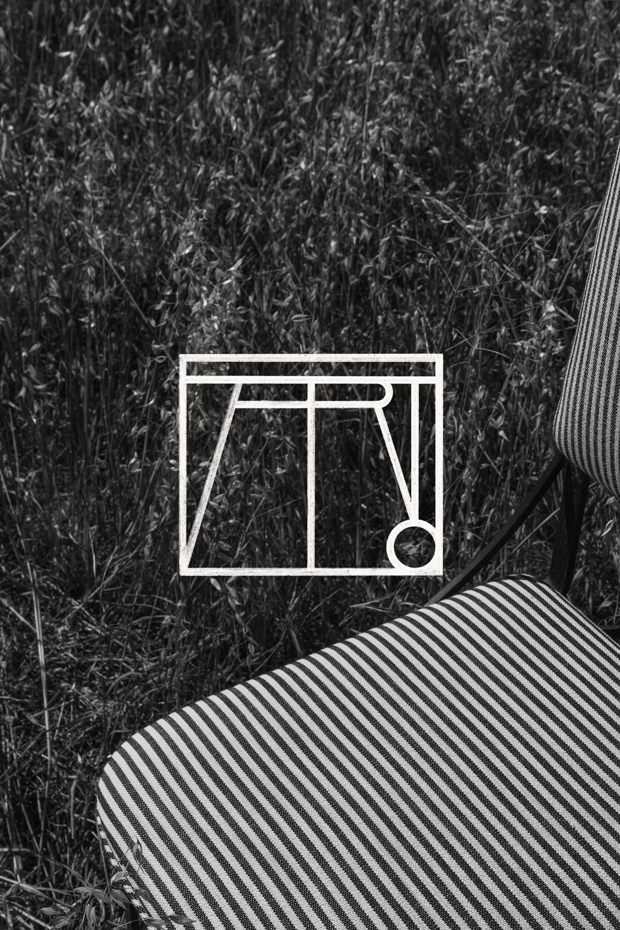Black and white photography of a striped chair in a grassy field, with a white graphic overlay of a geometric design.  
