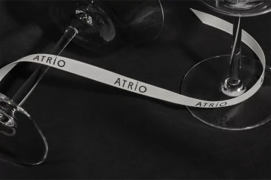 Branded ribbon reading ATRIO woven between two crystal wine glasses resting on black fabric