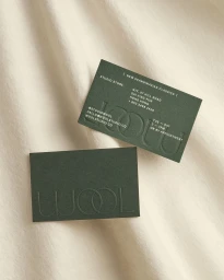 Two dark green business cards featuring debossed typography resting on a cream-coloured fabric background.