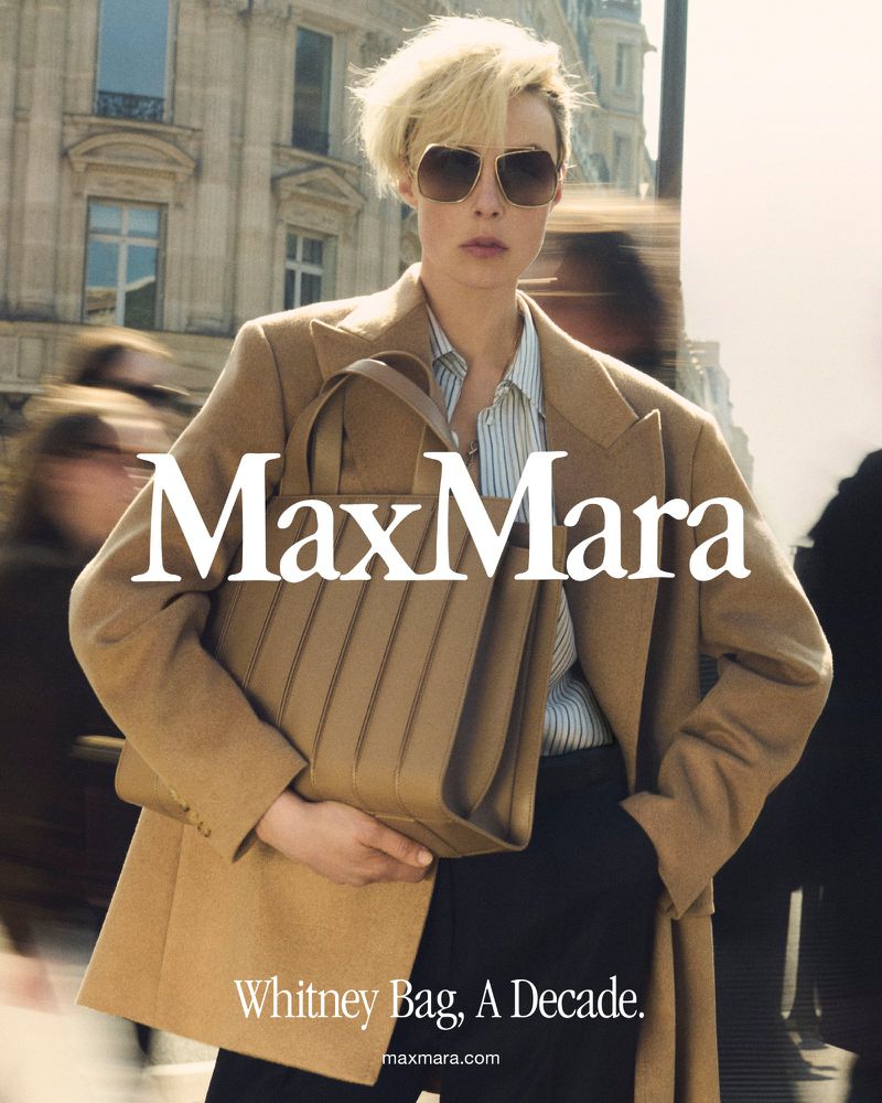 Fashion campaign featuring a blonde adult woman wearing a tan coat and sunglasses, holding a tan handbag, with a city street background.