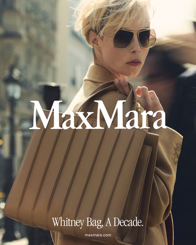 A woman with short blond hair wearing sunglasses, a beige coat, and carrying a Max Mara Whitney bag stands on a city street.