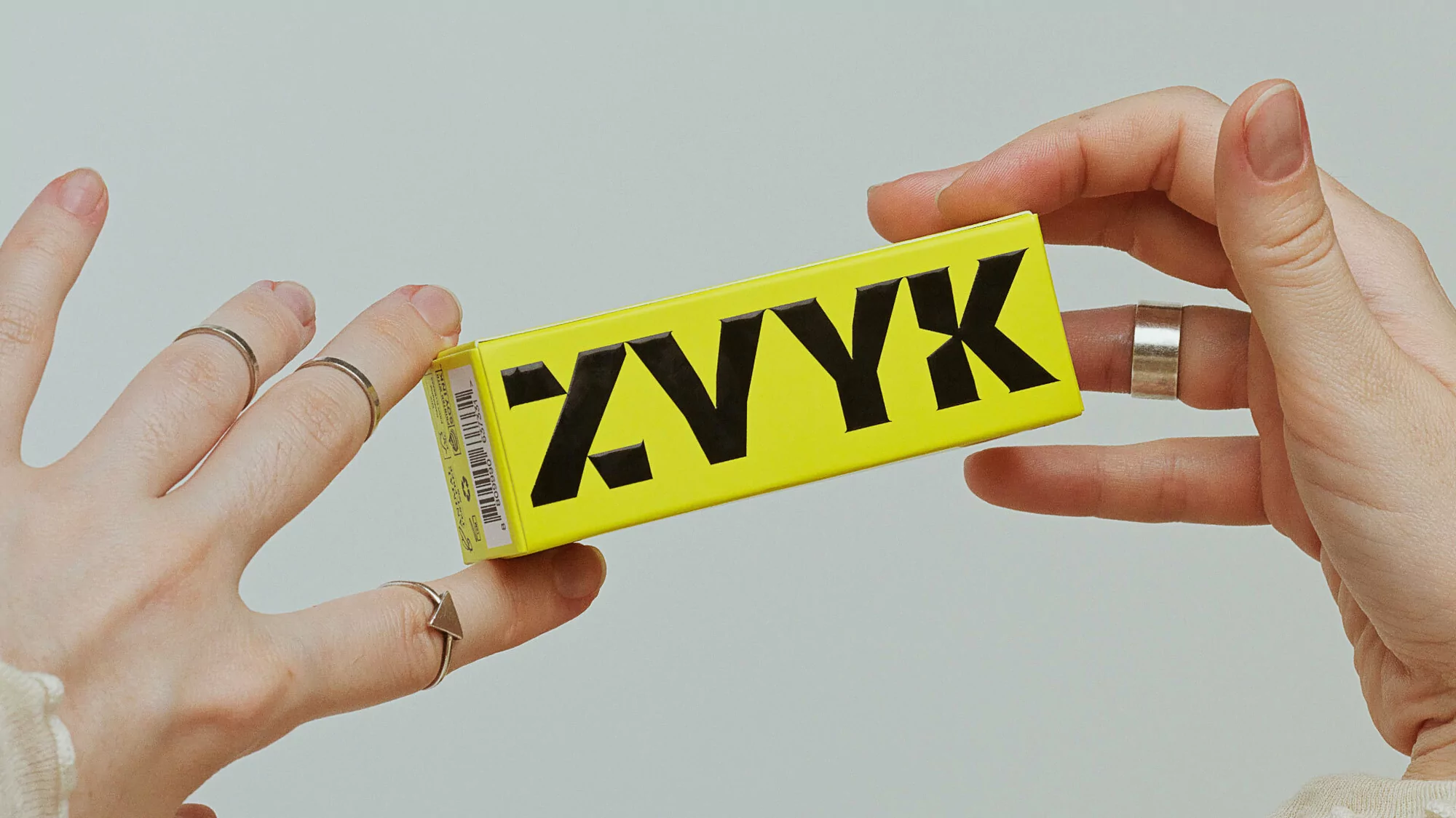 Hands hold a yellow box with the text 'ZYVX' in black. The background is neutral, highlighting the product and hands.