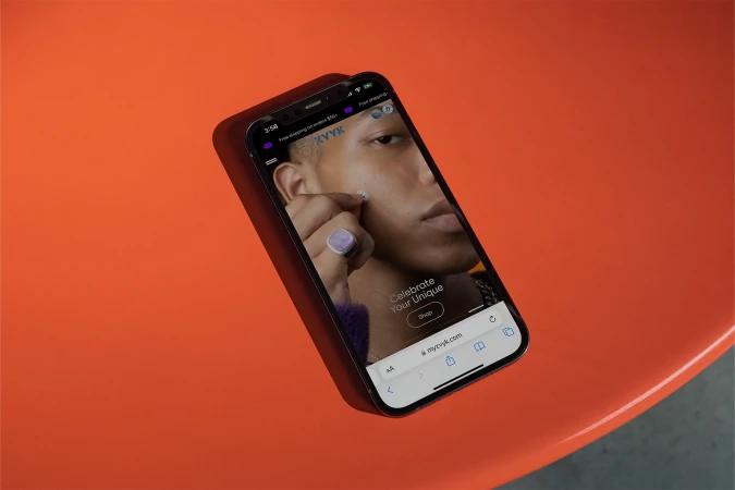 Close-up of a black smartphone displaying the myvzyk.com e-commerce website with a model wearing a ring, on an orange surface.
