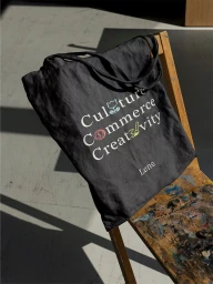 Dark grey canvas tote bag with white typography resting on a rustic wooden artist chair on a concrete floor.