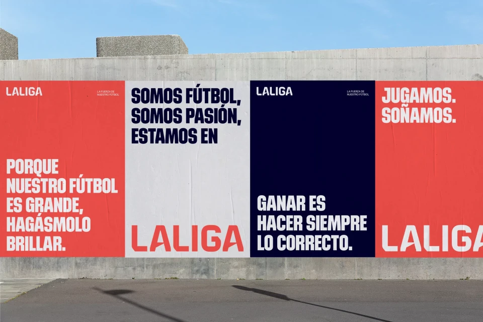 Outdoor view of La Liga posters on a concrete wall. The posters contain text in Spanish, with a palette of red, white, and dark blue.