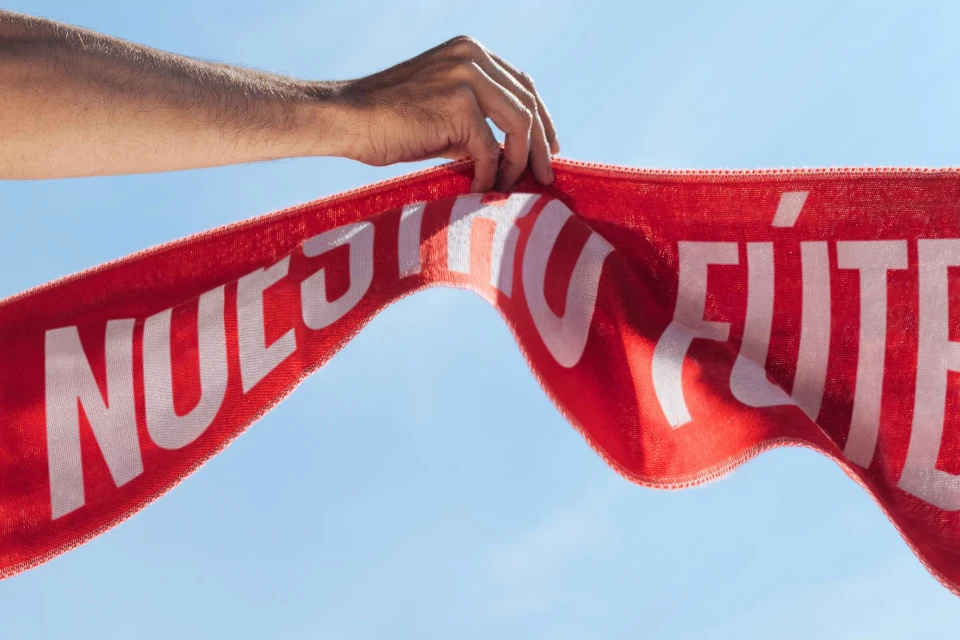 Arm holding red banner with white text 'Nuestro Futbol' against a blue sky background. Sports, fan, and patriotism themes.