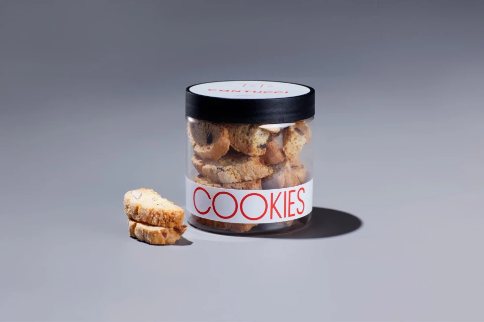 A jar filled with biscotti-style cookies sits beside two stacked biscotti on a grey surface, against a grey background, well-lit.
