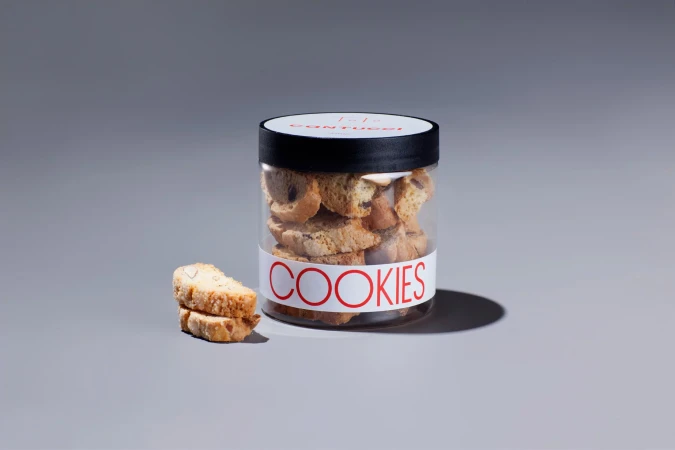 A jar filled with biscotti-style cookies sits beside two stacked biscotti on a grey surface, against a grey background, well-lit.