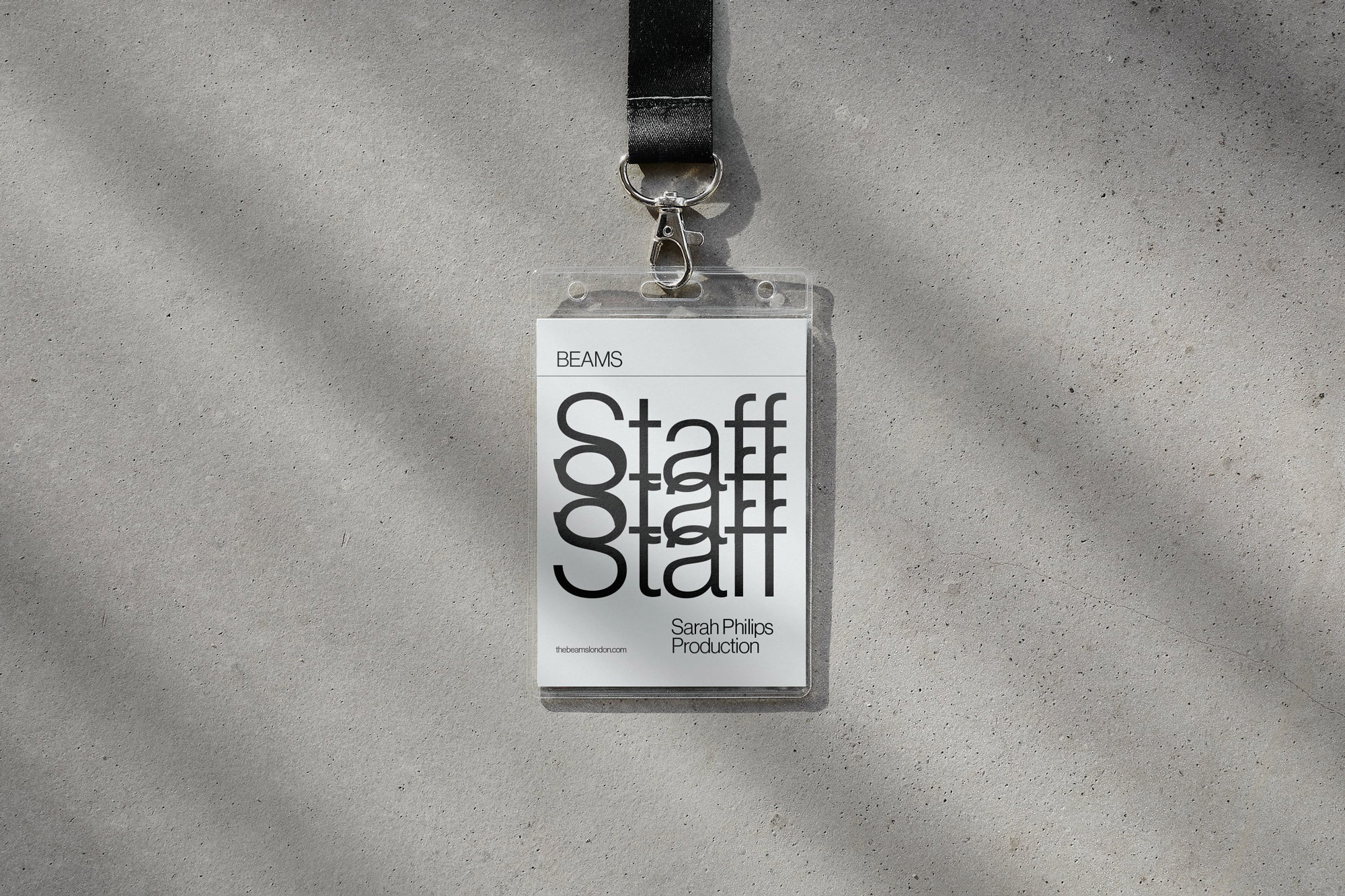 Staff badge by Beams hangs against a concrete wall with shadows. The badge is on a black lanyard and features white and black text.