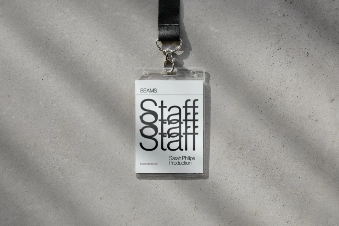 Staff badge by Beams hangs against a concrete wall with shadows. The badge is on a black lanyard and features white and black text.