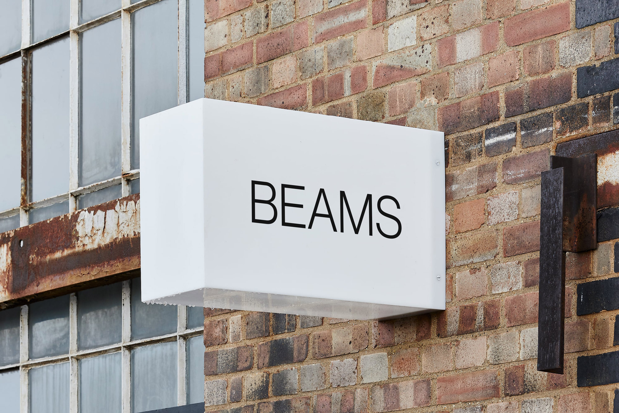 Exterior of a building featuring a white rectangular sign with the brand name BEAMS in black lettering mounted on a brick wall.