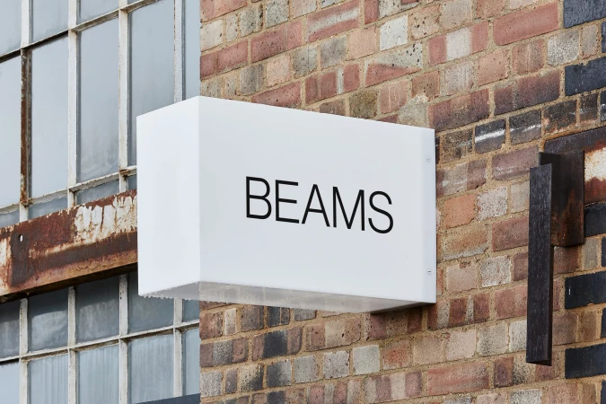 Exterior of a building featuring a white rectangular sign with the brand name BEAMS in black lettering mounted on a brick wall.