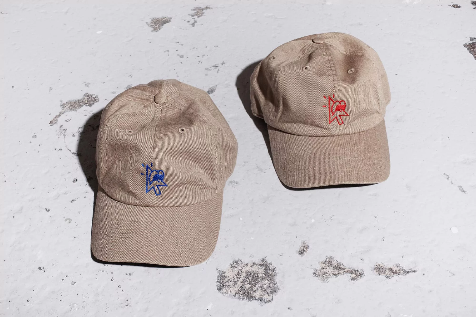 Two tan baseball caps sit on a concrete surface, each with a unique, colorful graphic embroidered on the front panel.