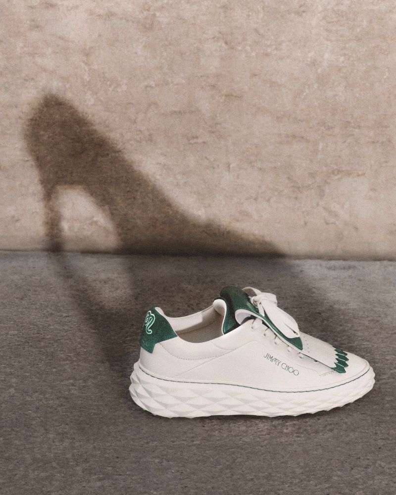 White leather sneaker with green accents, laces, and textured sole with a high-heel shoe shadow on a neutral textured wall.