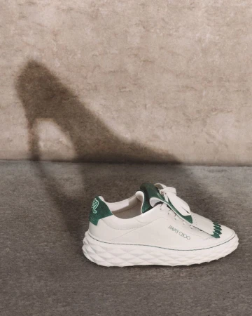 White leather sneaker with green accents, laces, and textured sole with a high-heel shoe shadow on a neutral textured wall.