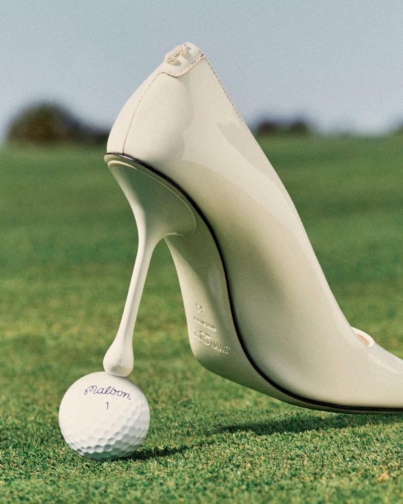 Elegant cream-colored stiletto heel balances on a golf ball with the green grass and soft-focus trees in the background.