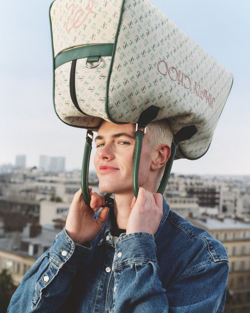 Young adult male poses with a beige Jimmy Choo duffel bag atop his head, wearing a blue denim jacket against an urban cityscape.