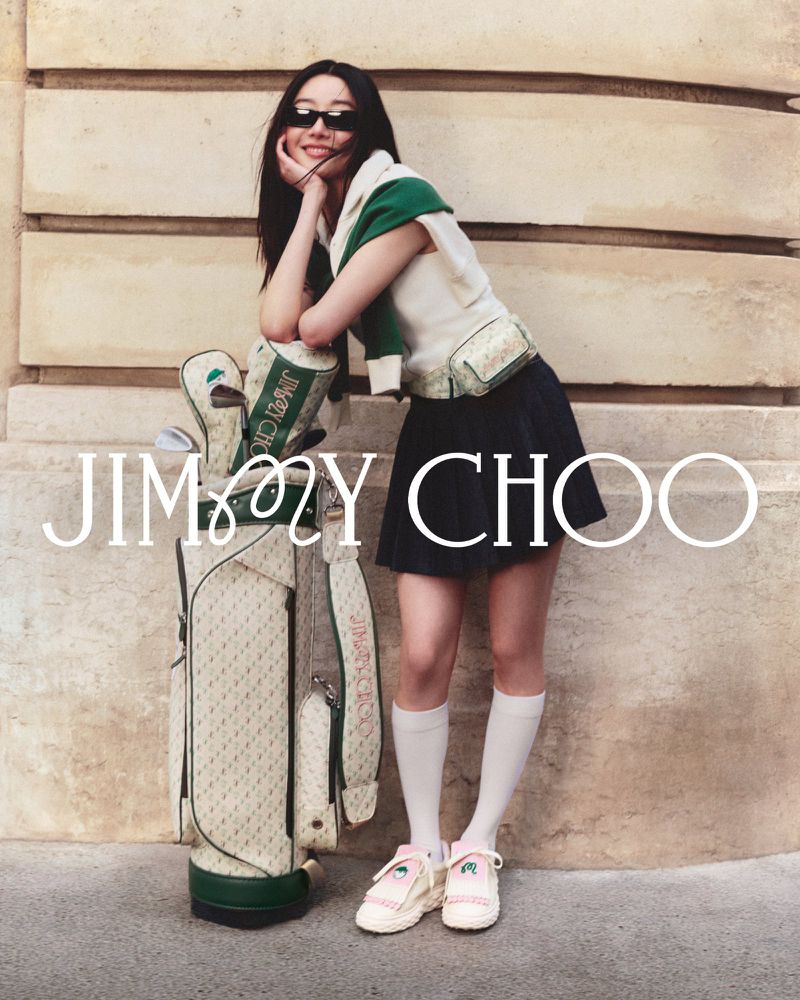 A woman in a preppy outfit leans against a golf bag with the Jimmy Choo logo, against a neutral wall outdoors in a fashion lifestyle shot.