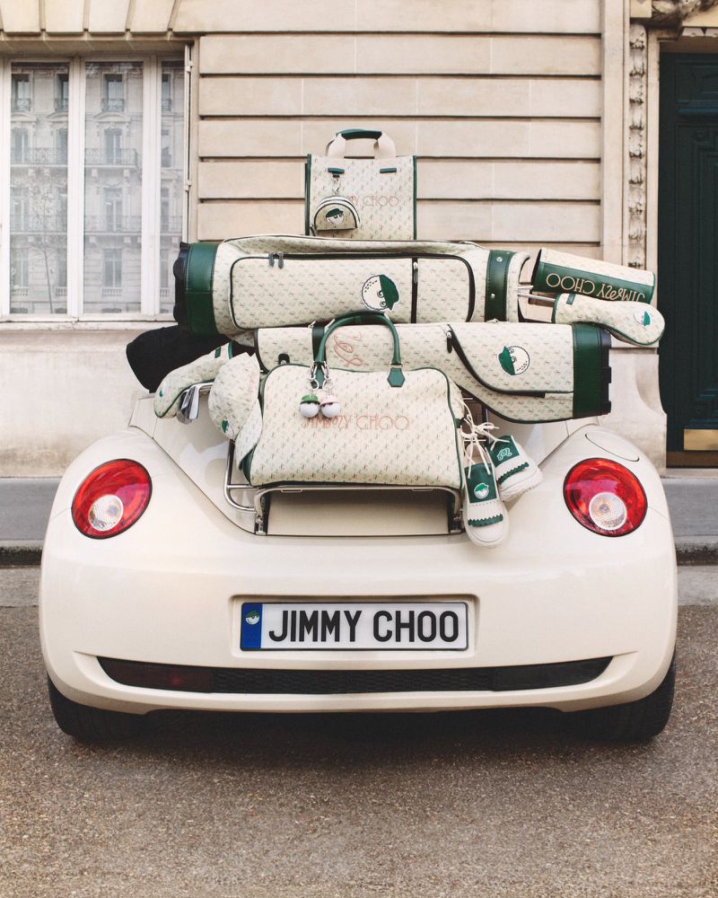 Jimmy Choo branded golf bags and accessories are stacked on the back of a vintage car with a building in the background.