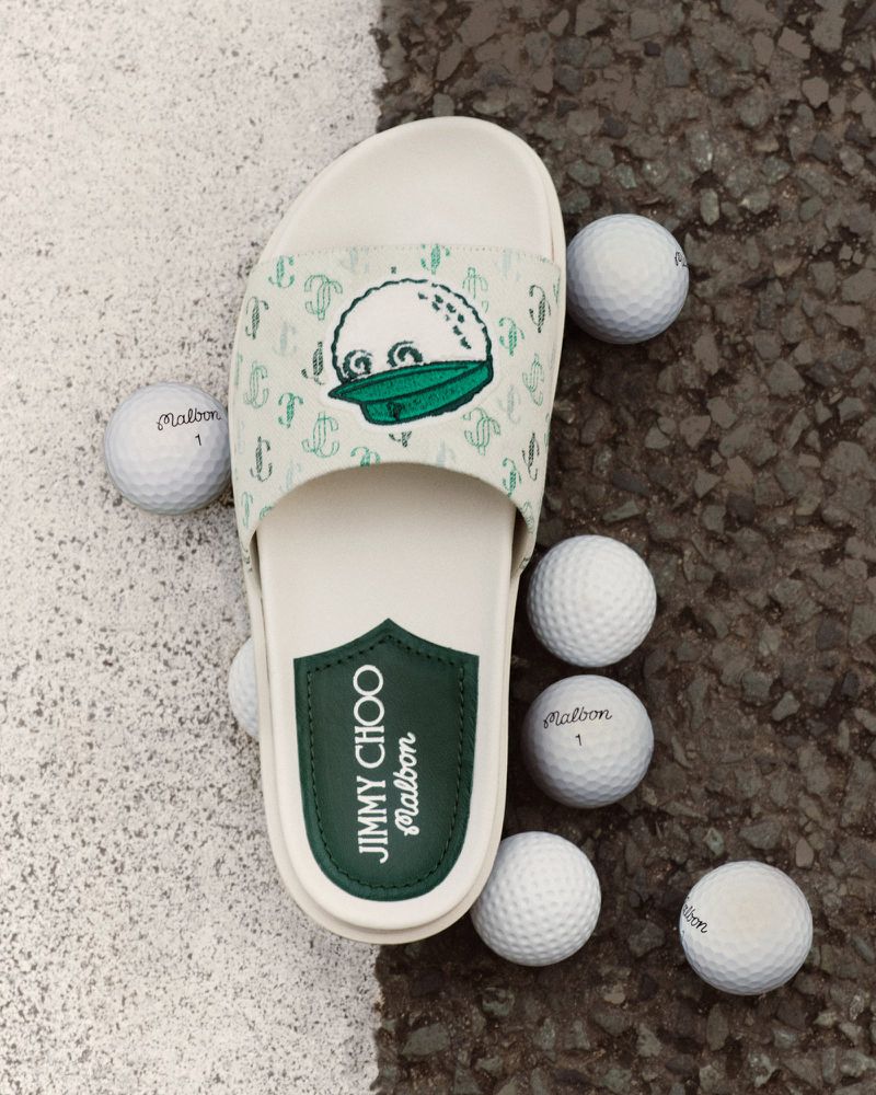 Jimmy Choo Malbon slide sandal patterned with a golf ball character, surrounded by white golf balls, placed on sand and asphalt.