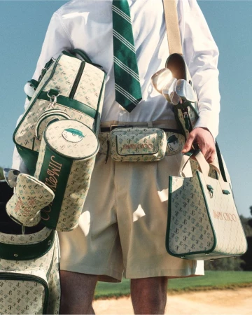 A man in golf attire, including a shirt, shorts, and tie, is carrying several patterned golf bags and a tote bag in an outdoor setting.