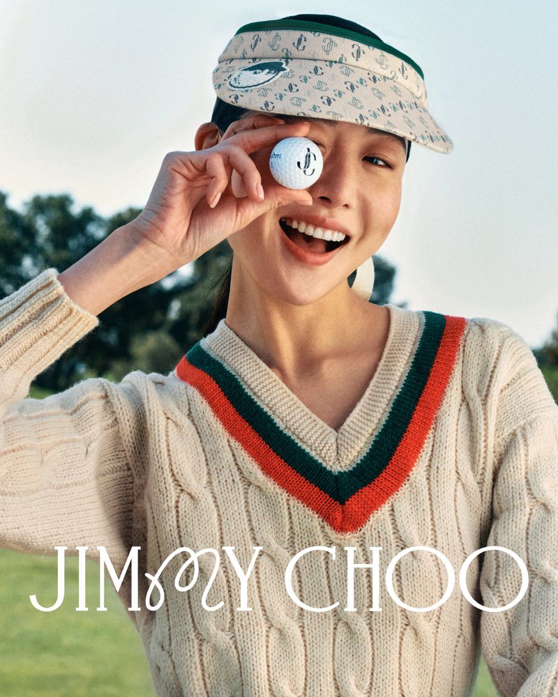 Woman in a visor and cricket sweater smiles holding a golf ball near her eye with the Jimmy Choo logo in a promotional image.