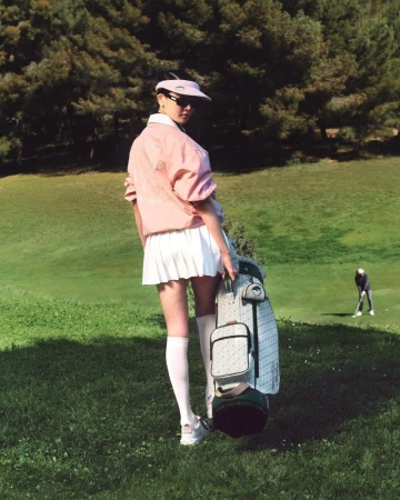 A woman in golf attire carries a golf bag on a green golf course on a sunny day with trees in the background as another person plays.