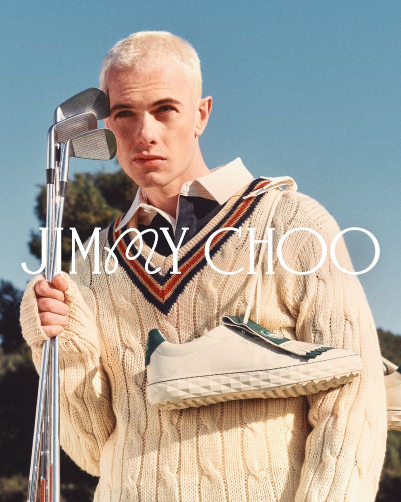 Man in a cricket sweater holds golf clubs and a Jimmy Choo shoe, set against a blue sky and trees. Fashion advertising campaign.