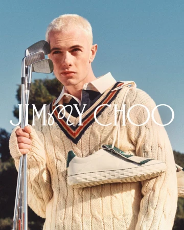 Man in a cricket sweater holds golf clubs and a Jimmy Choo shoe, set against a blue sky and trees. Fashion advertising campaign.