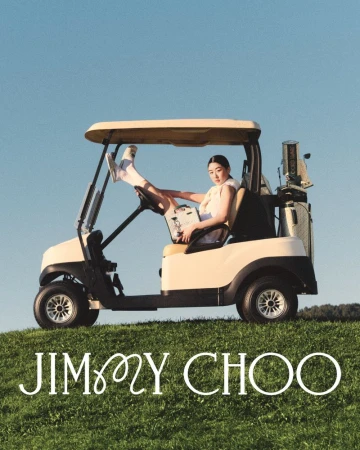 Woman in golf cart holding purse, wearing sneakers and socks with Jimmy Choo logo on green grass and a blue sky.