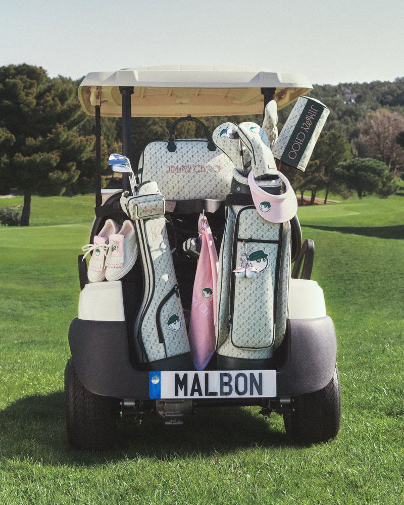 Golf cart filled with golf bags and accessories, featuring a Jimmy Choo handbag and Malbon license plate, set on a lush green golf course.